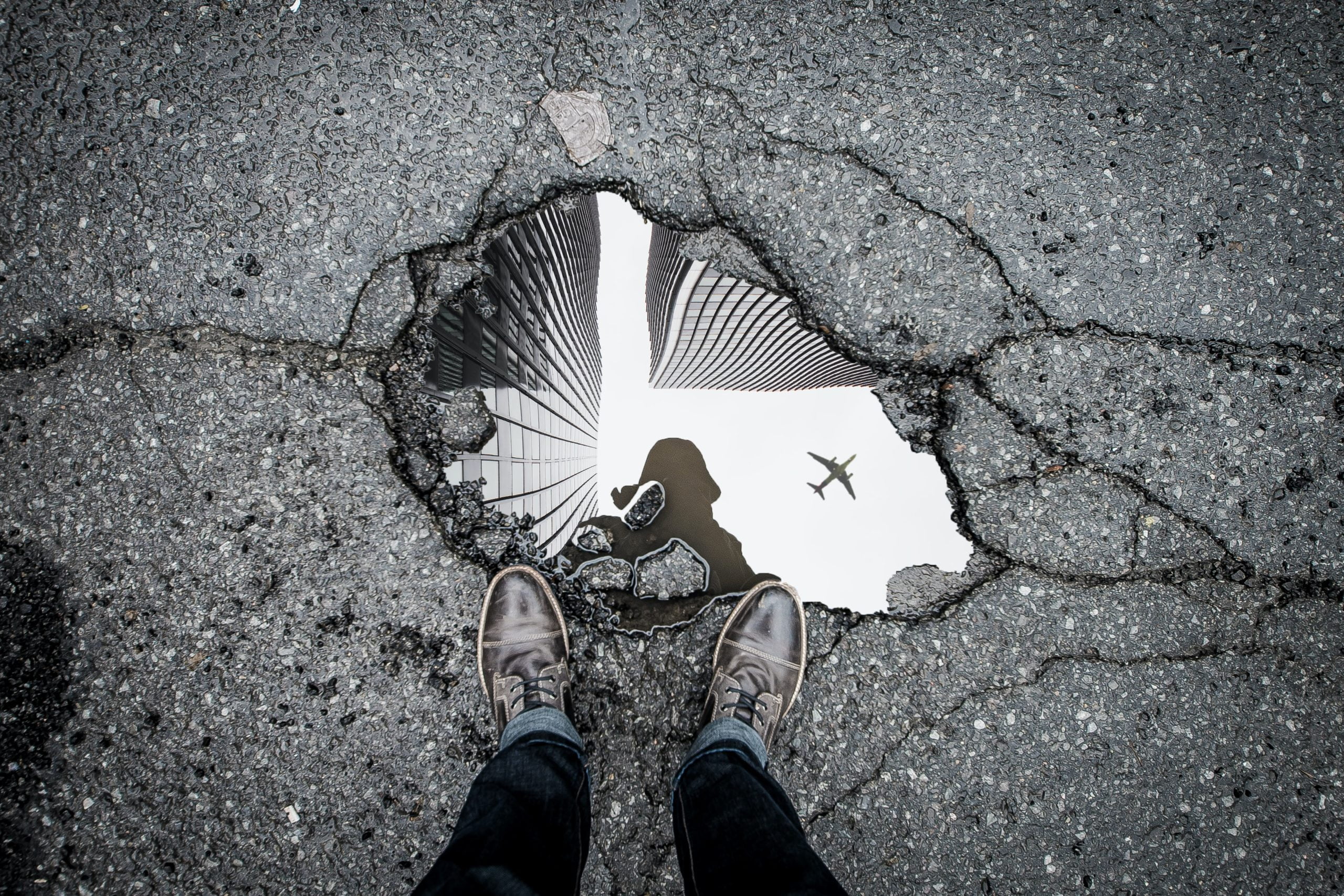 marc-olivier-jodoin–TQUERQGUZ8-unsplash Person's shoes by a puddle reflecting skyscrapers, a plane, and the person's silhouette on cracked asphalt.