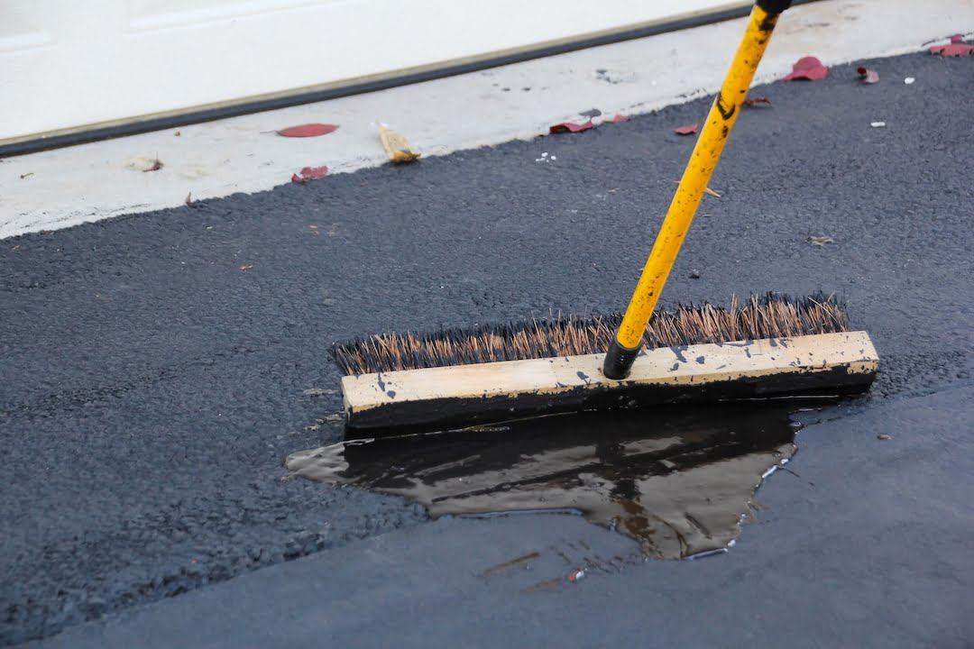 A broom sweeps water off a black asphalt driveway near a white garage door.