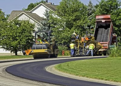 Workers paving a curved road near houses with construction vehicles and equipment on a sunny day.