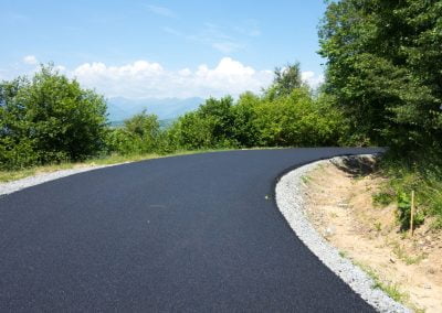 A newly paved asphalt road curves through a green, wooded area under a blue sky.