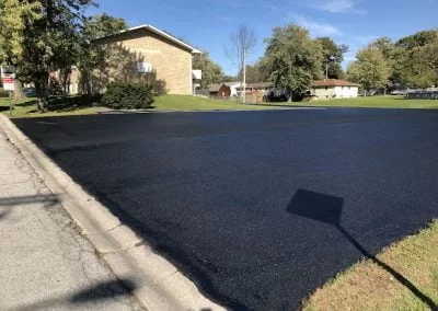 Asphalt Pavement IMG_3042-400×284-1 Freshly paved asphalt pavement parking lot next to a sidewalk and residential buildings on a sunny day.