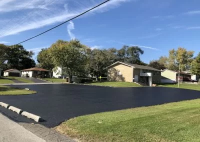 Freshly paved asphalt driveway with professional sealcoating in front of suburban houses on a sunny day, surrounded by green grass and trees.