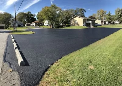 Paving Contractors Freshly paved black asphalt driveway, crafted by expert paving contractors, sits beside green grass and houses on a sunny day.