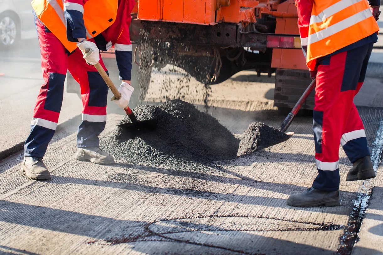 asphalt paving company: Asphalt Paving Jones Asphalt Workers in orange safety vests and red uniforms using tools to spread asphalt from a truck onto a road.