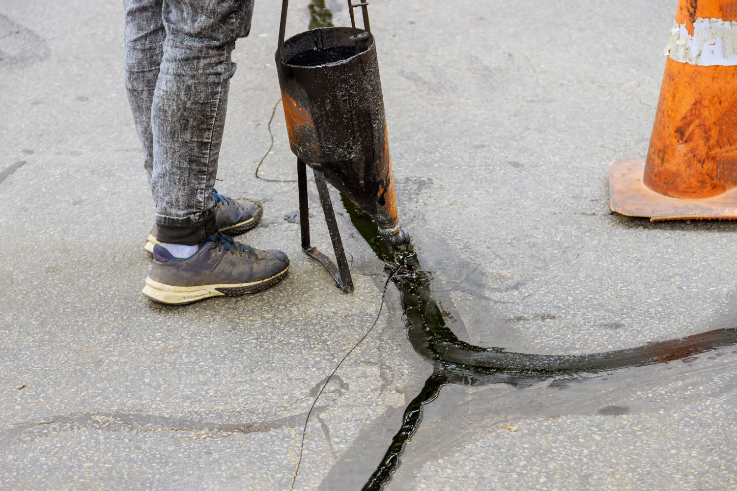 9106 Person sealing cracks in pavement with liquid asphalt beside an orange traffic cone, preparing the surface for Asphalt Driveway Resurfacing.