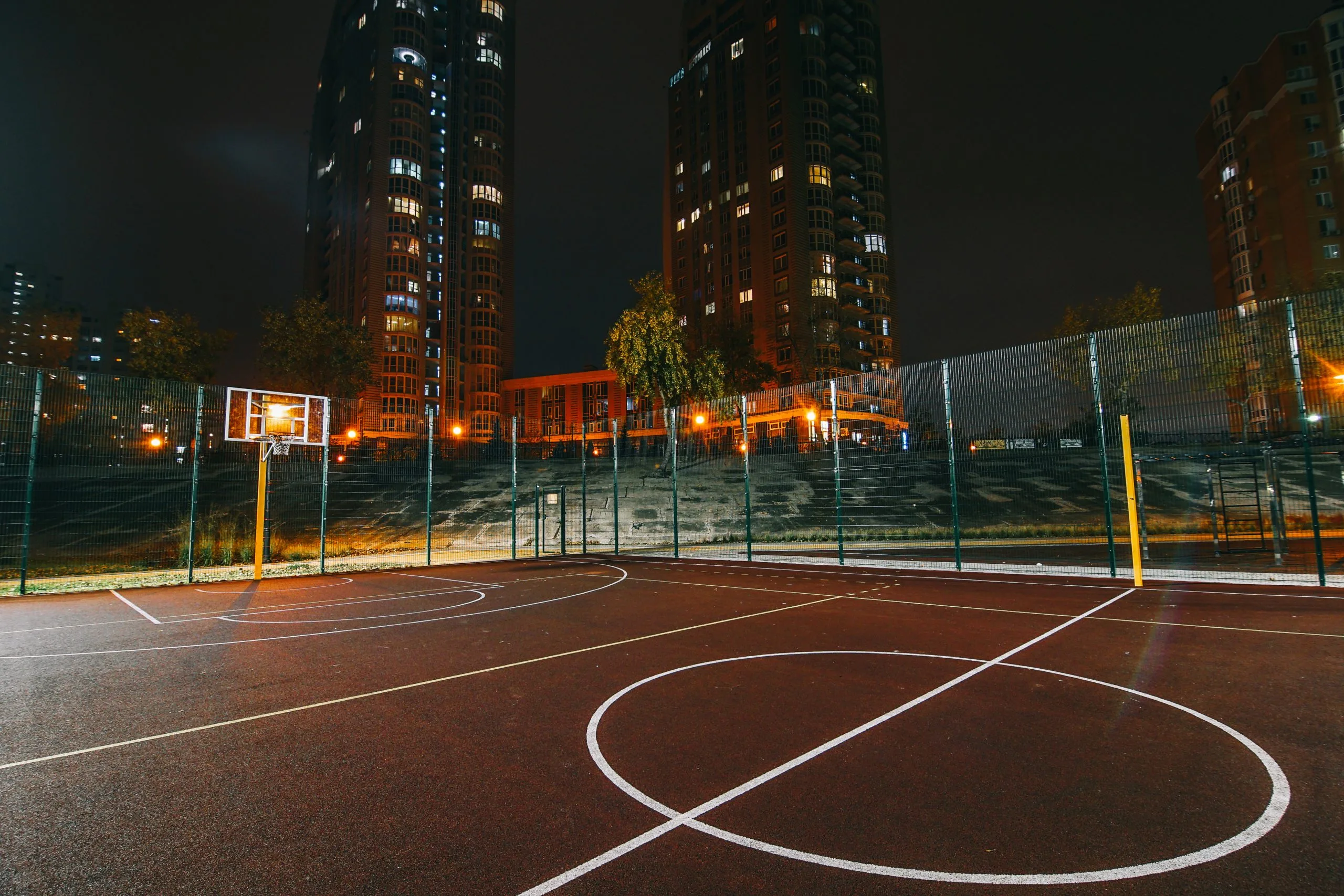 Sport Courts Paving A dimly lit outdoor basketball court at night with tall buildings in the background.