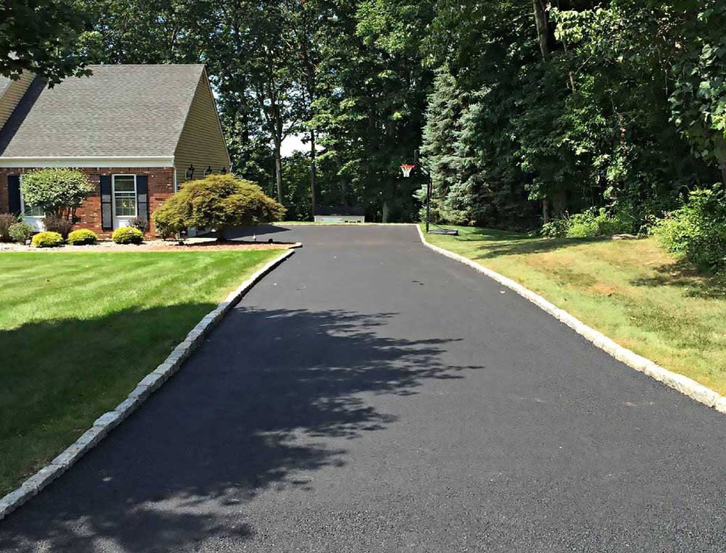 Freshly paved asphalt driveway leading to a house, bordered by grass and trees on a sunny day.