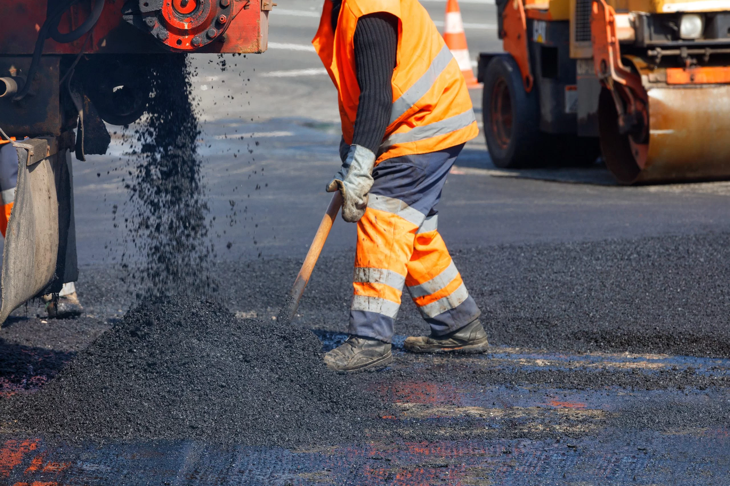 Worker in an orange safety vest and pants spreading asphalt on a road with construction machinery nearby.