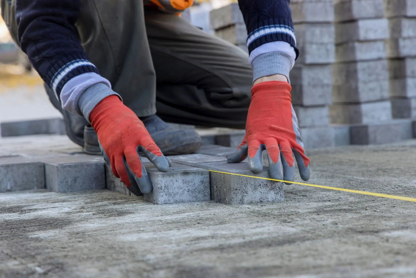 patio A worker with red gloves places paving stones and measures distance with a yellow tape on a construction site.