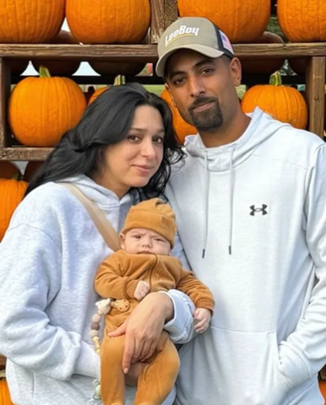 A couple holds a baby in front of a display of pumpkins, all wearing cozy fall outfits.