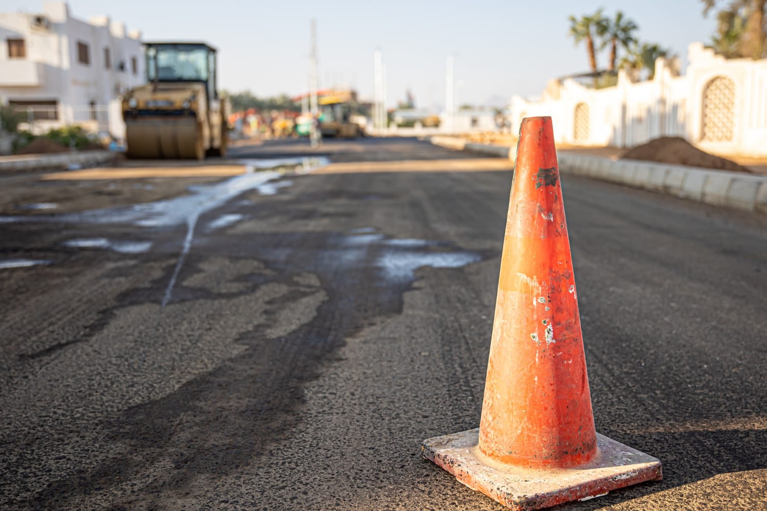An orange traffic cone on a newly paved road with construction equipment in the background.