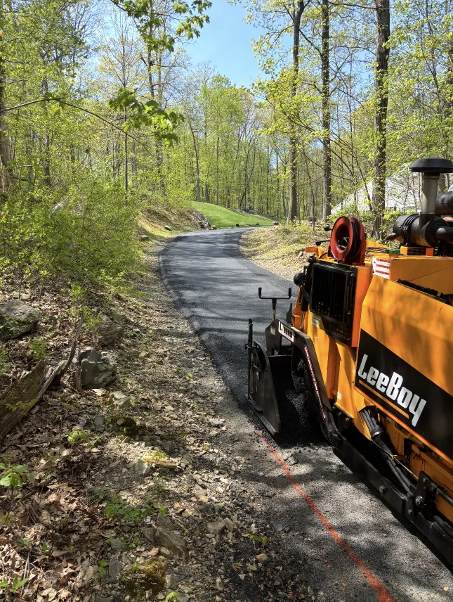 Yellow LeeBoy paving machine laying fresh asphalt on a winding road through a green, wooded area.