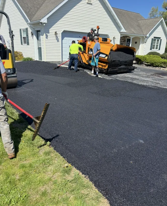 Workers paving a driveway with fresh asphalt using machinery and rakes in front of a white house.